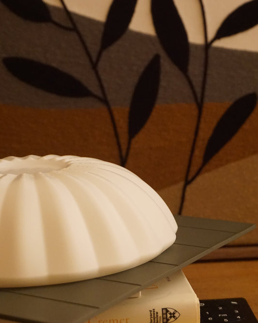 Close-up of the Kueh Tutu lamp's beautifully curved shade, resting on a stack of books, with a fan remote and felt painting visible in the low-angle background