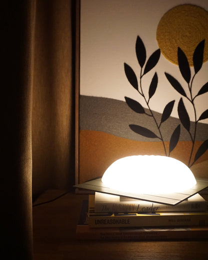 Illuminated Kueh Tutu lamp casting a beautiful ambient glow that highlights the nearby curtain and felt painting, with soft light spilling onto the books below