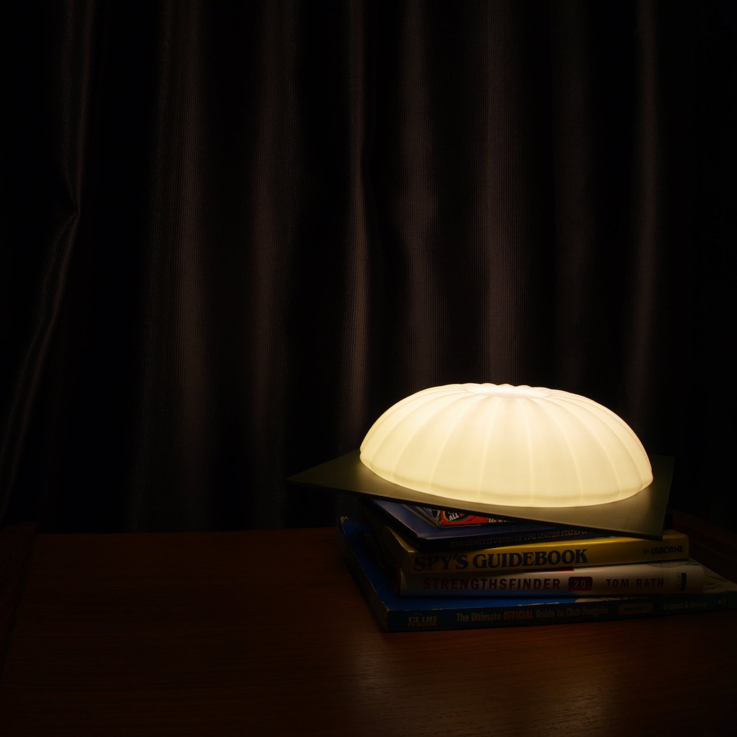 Illuminated Kueh Tutu lamp casting a warm ambient glow on a curtain background, with soft light spilling onto a stack of books below