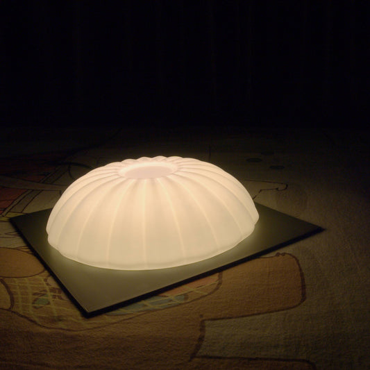 Illuminated Kueh Tutu lamp glowing in complete darkness while resting on a bed, highlighting its soft ambient light