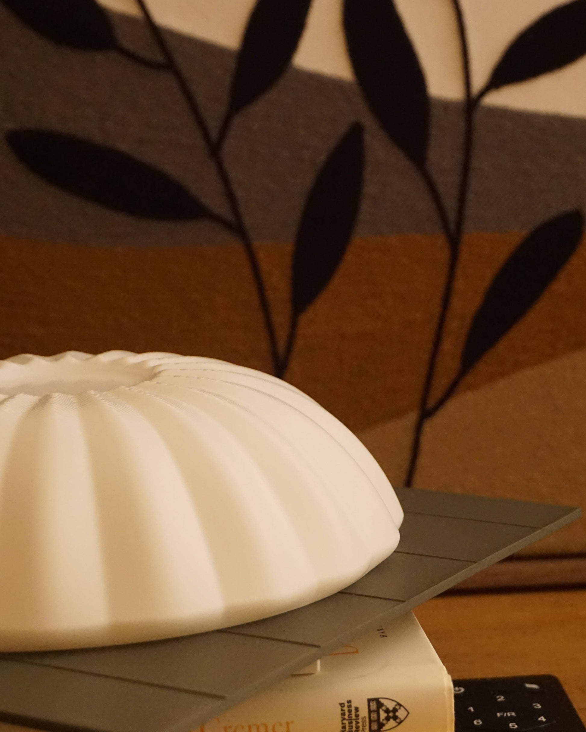 Close-up of the Kueh Tutu lamp's beautifully curved shade, resting on a stack of books, with a fan remote and felt painting visible in the low-angle background
