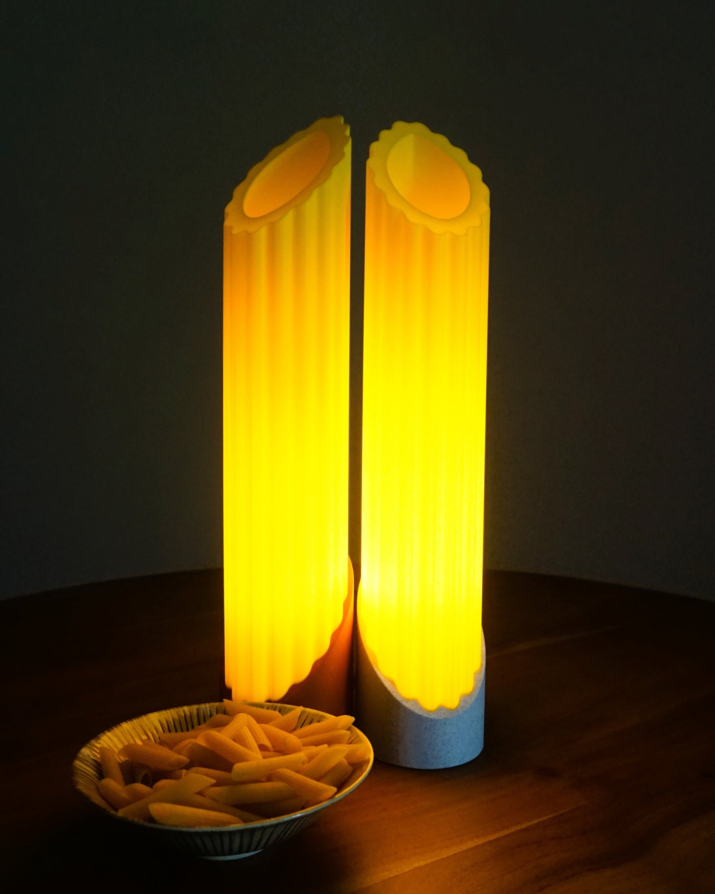 Two Penne pasta lamps glowing warmly in the dark with a bowl of uncooked penne pasta