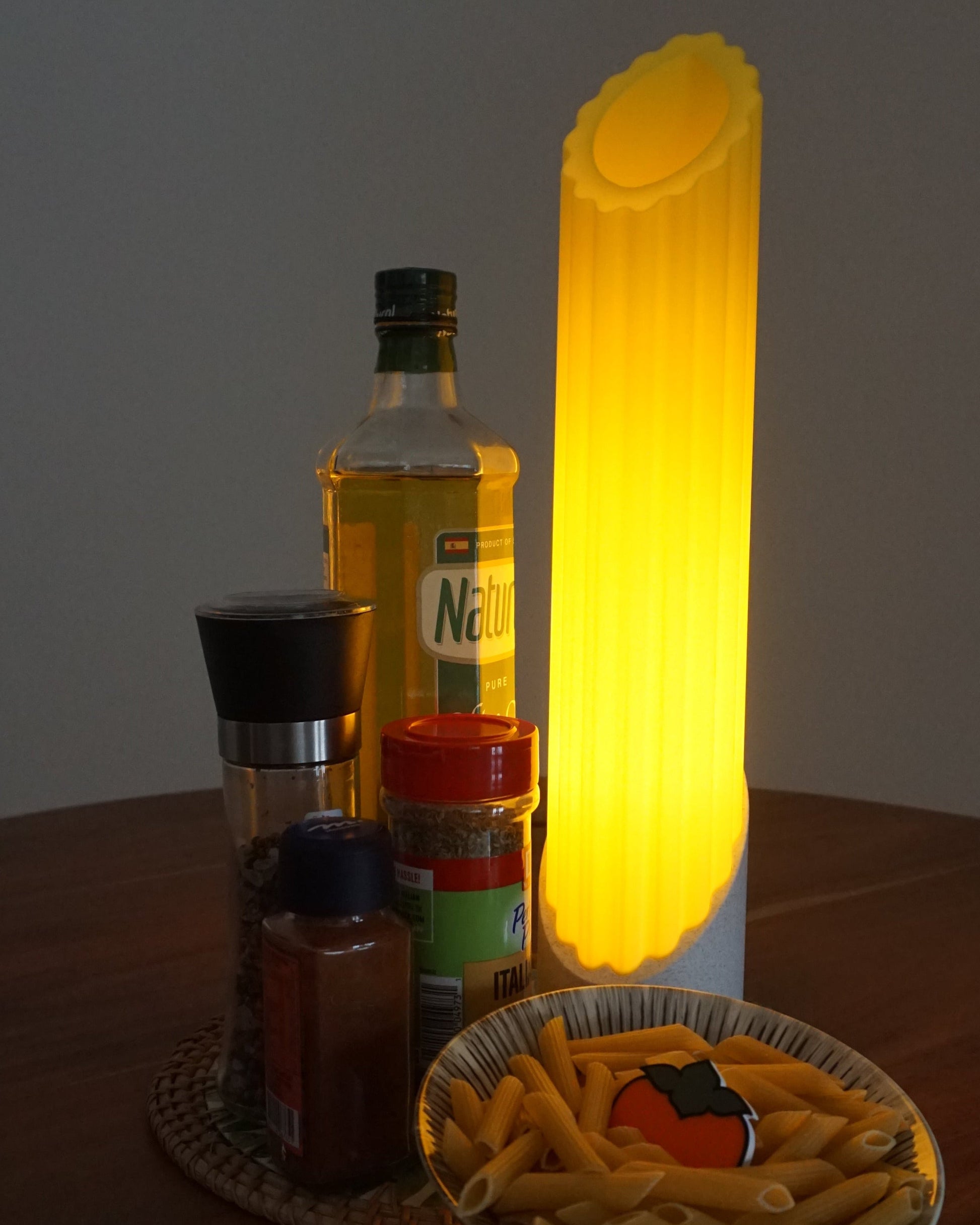 Single Penne pasta lamp standing next to seasoning and a bowl of raw pasta, on top a mini persimmon logo