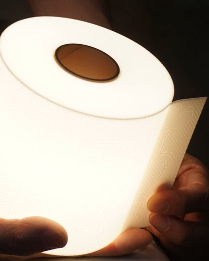 Close-up view of the illuminated toilet paper lamp, held at the base, angled to clearly show the top design and the light highlighting the central paper roll