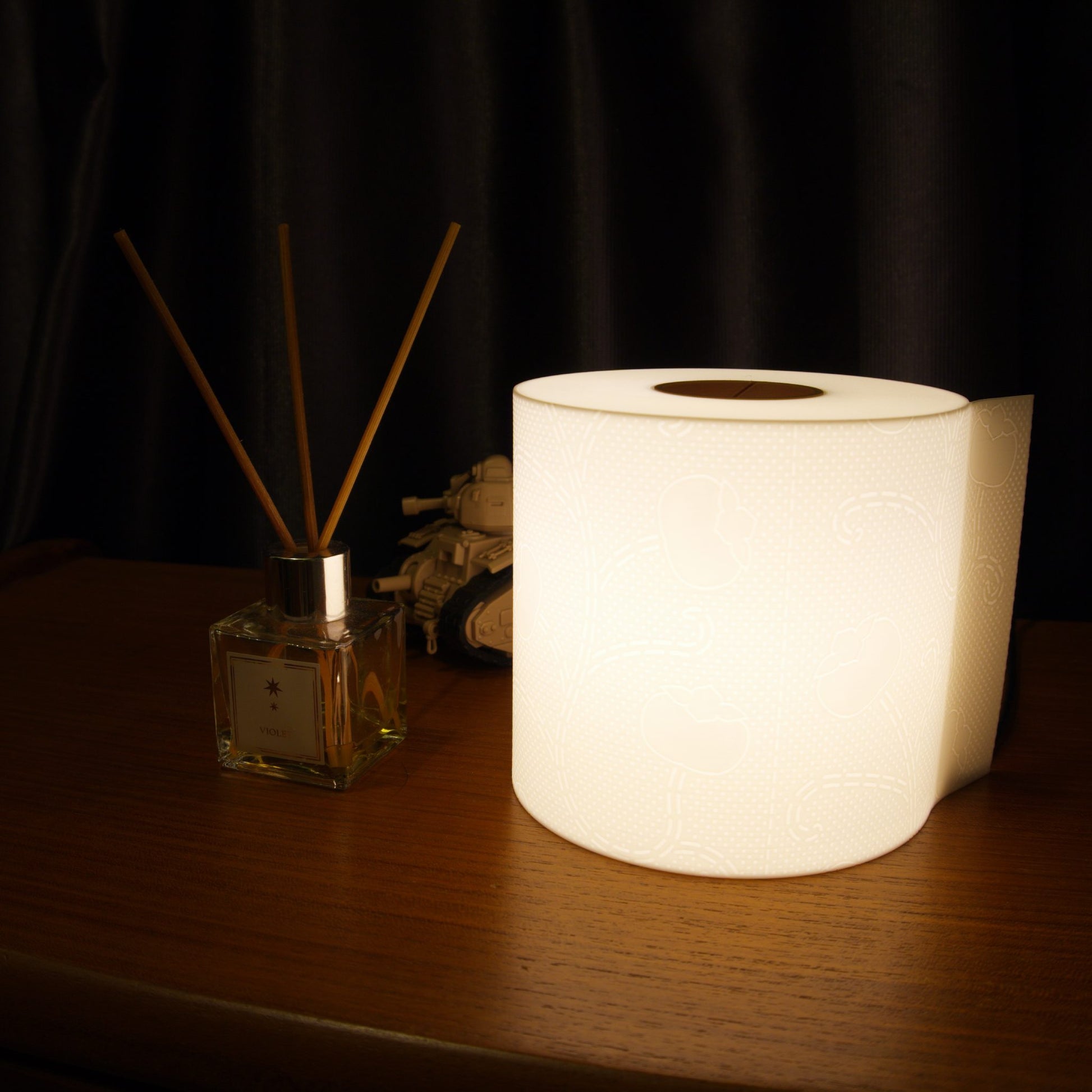 Illuminated toilet paper lamp on a bedside table surrounded by various personal items, casting a warm ambient glow