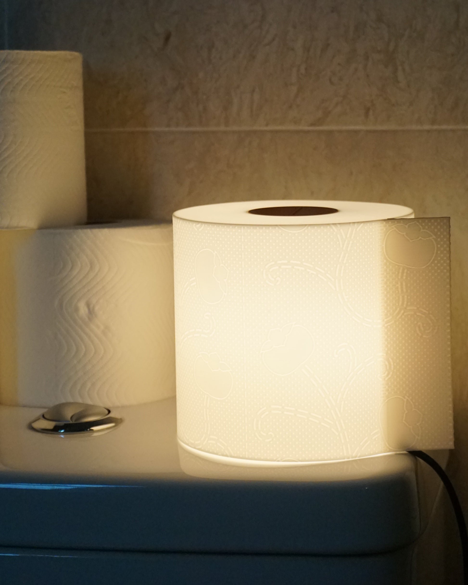 Illuminated toilet paper lamp casting a warm glow, placed on the toilet tank next to several rolls of real toilet paper