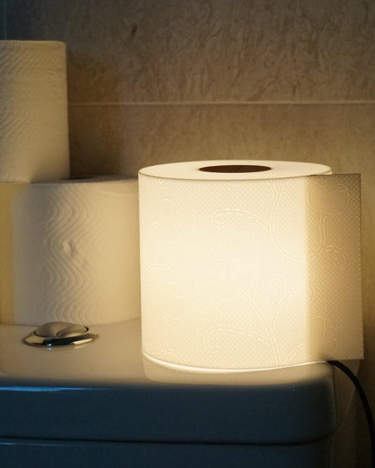 Illuminated toilet paper lamp casting a warm glow, placed on the toilet tank next to several rolls of real toilet paper