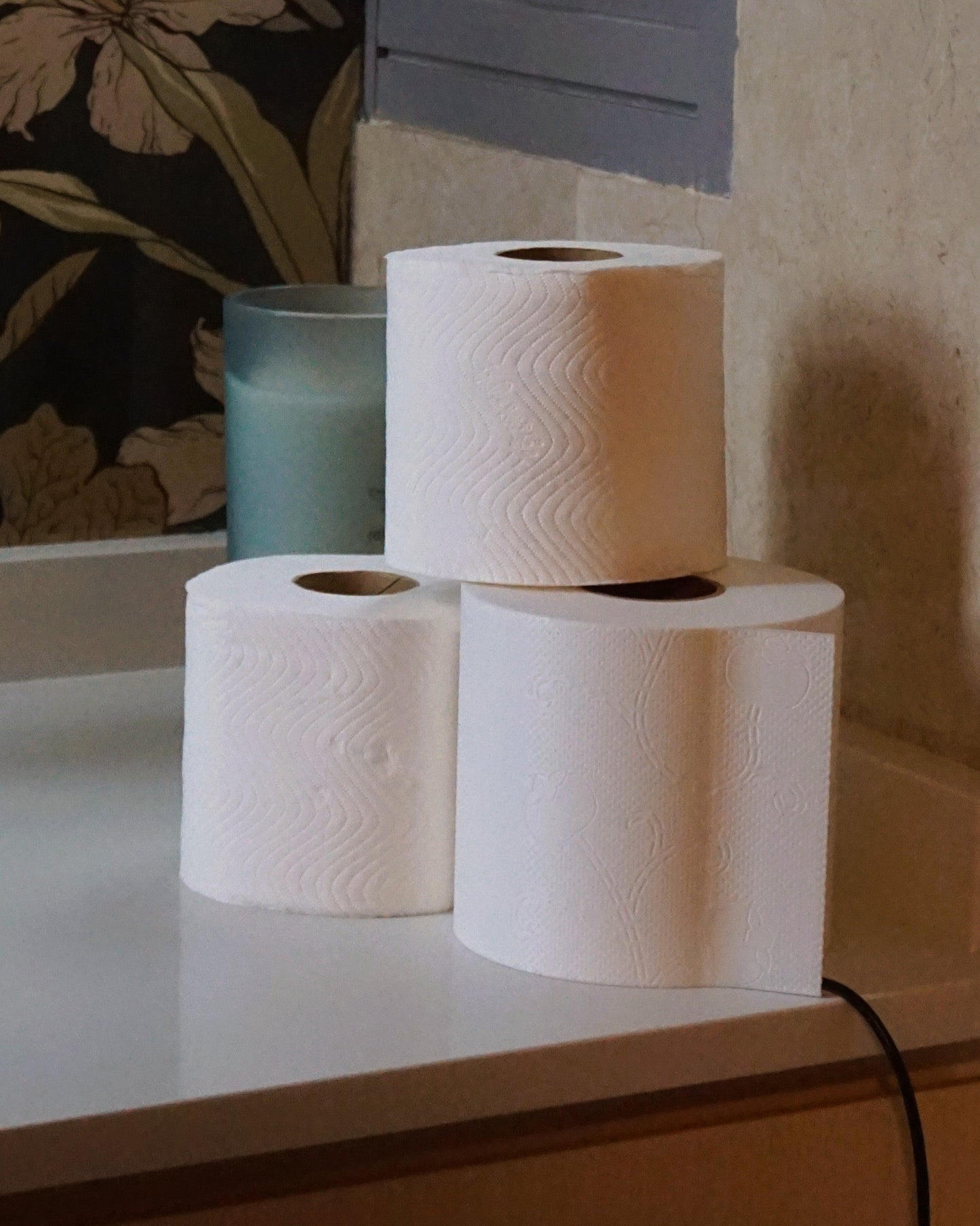 Unlit toilet paper lamp in bright daylight, creatively placed next to and stacked on real toilet paper rolls for thematic styling