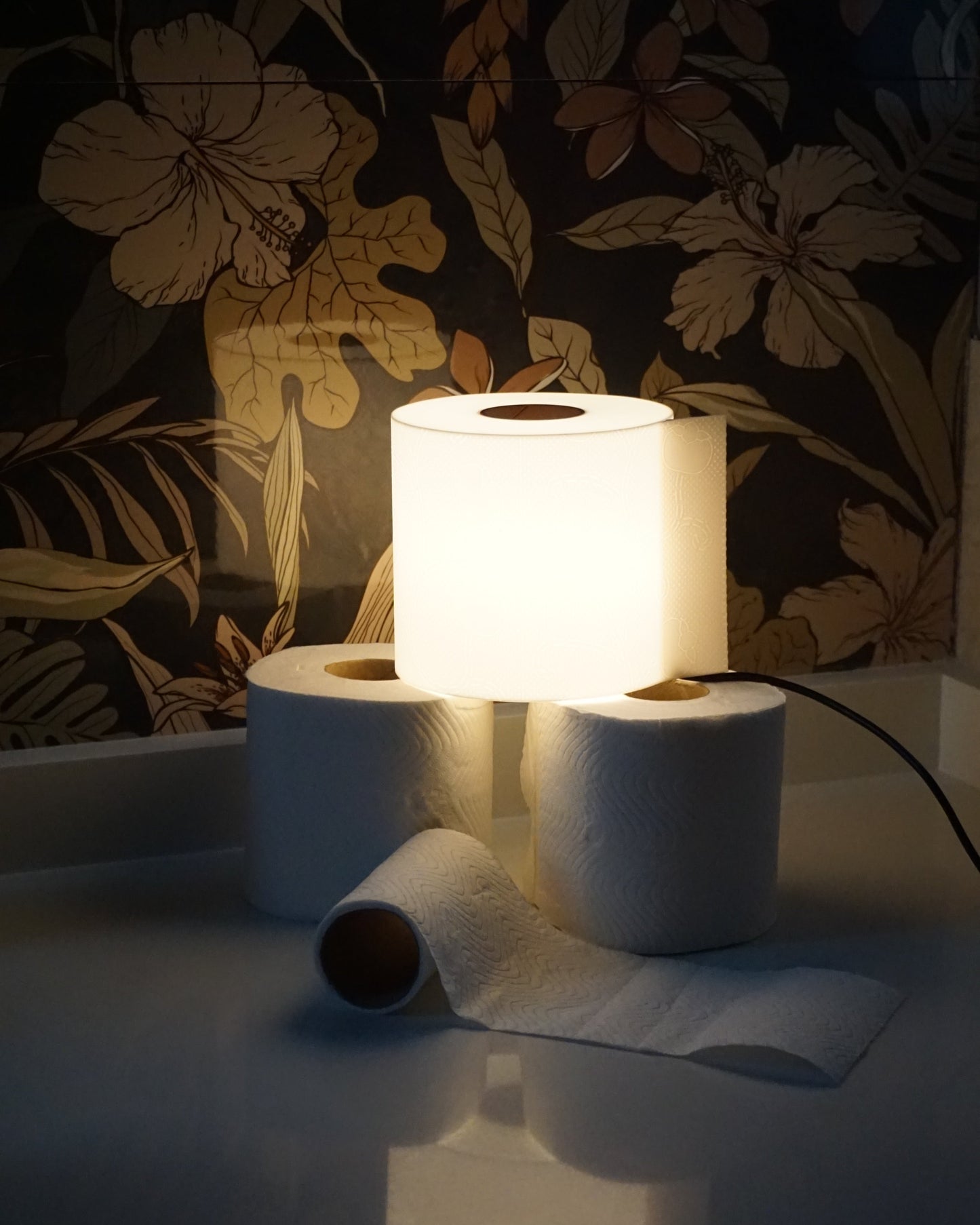 Illuminated toilet paper lamp resting on two stacked toilet rolls, casting a gentle ambient glow onto the flowery wall